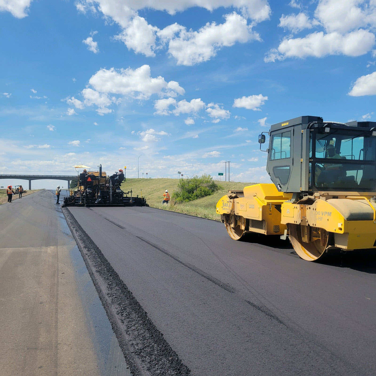 Construction workers operating machinery to lay asphalt on a highway under a blue sky with clouds.