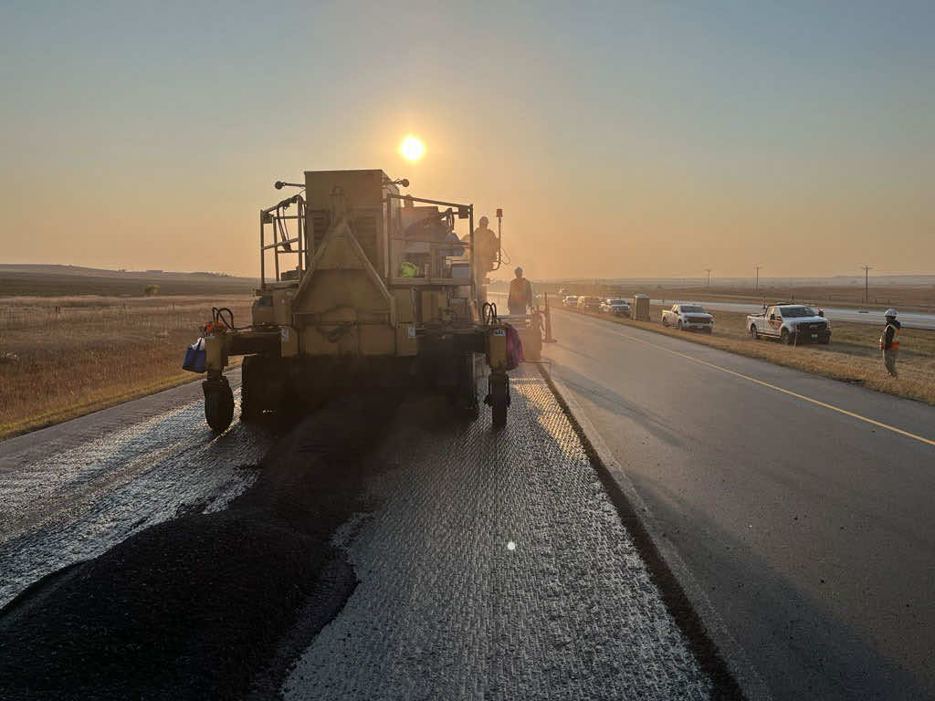 Construction workers operating a paving machine on a highway at sunset.