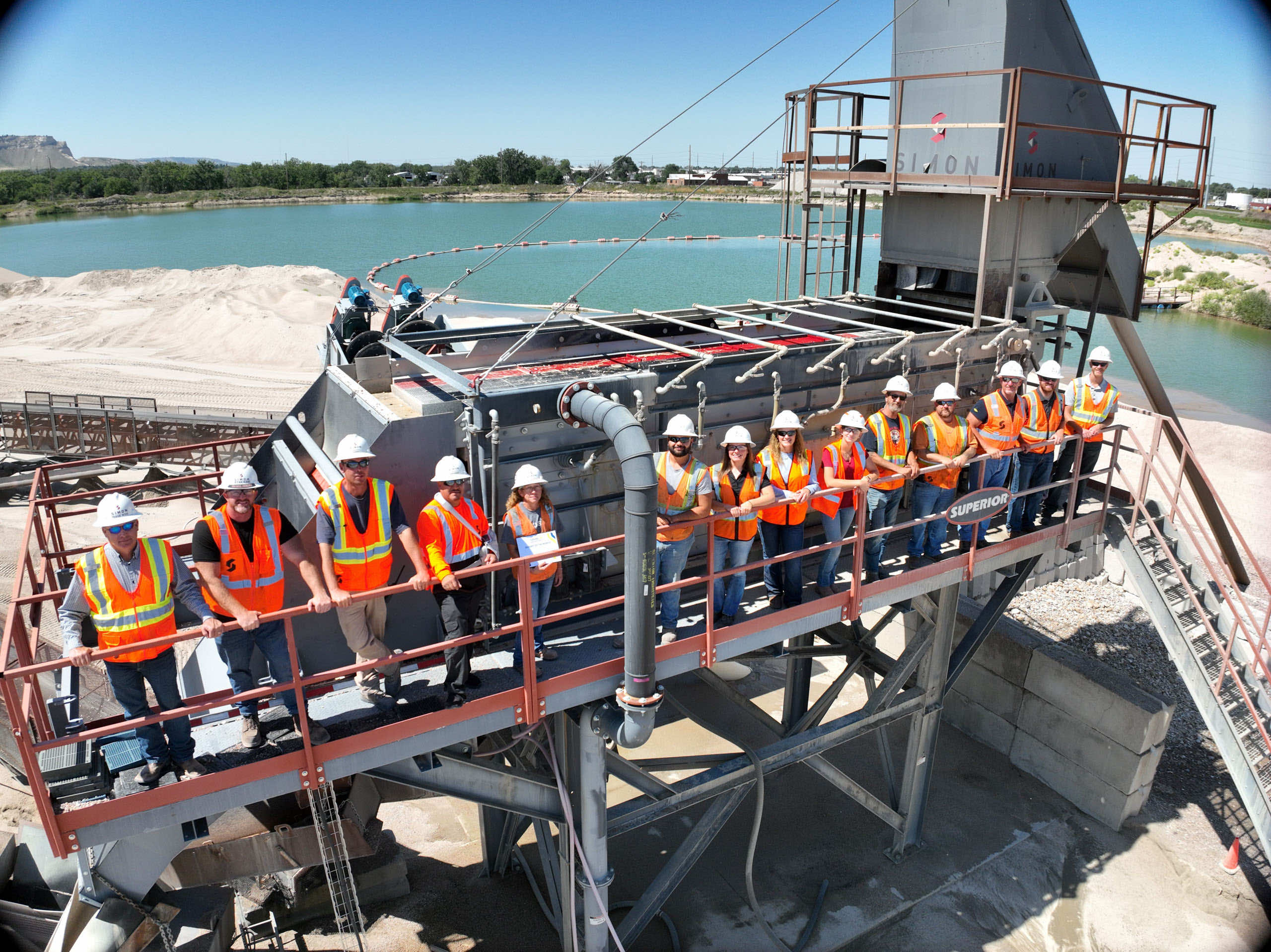 A group of workers in safety vests and helmets standing on a platform at a manufacturing site near water.