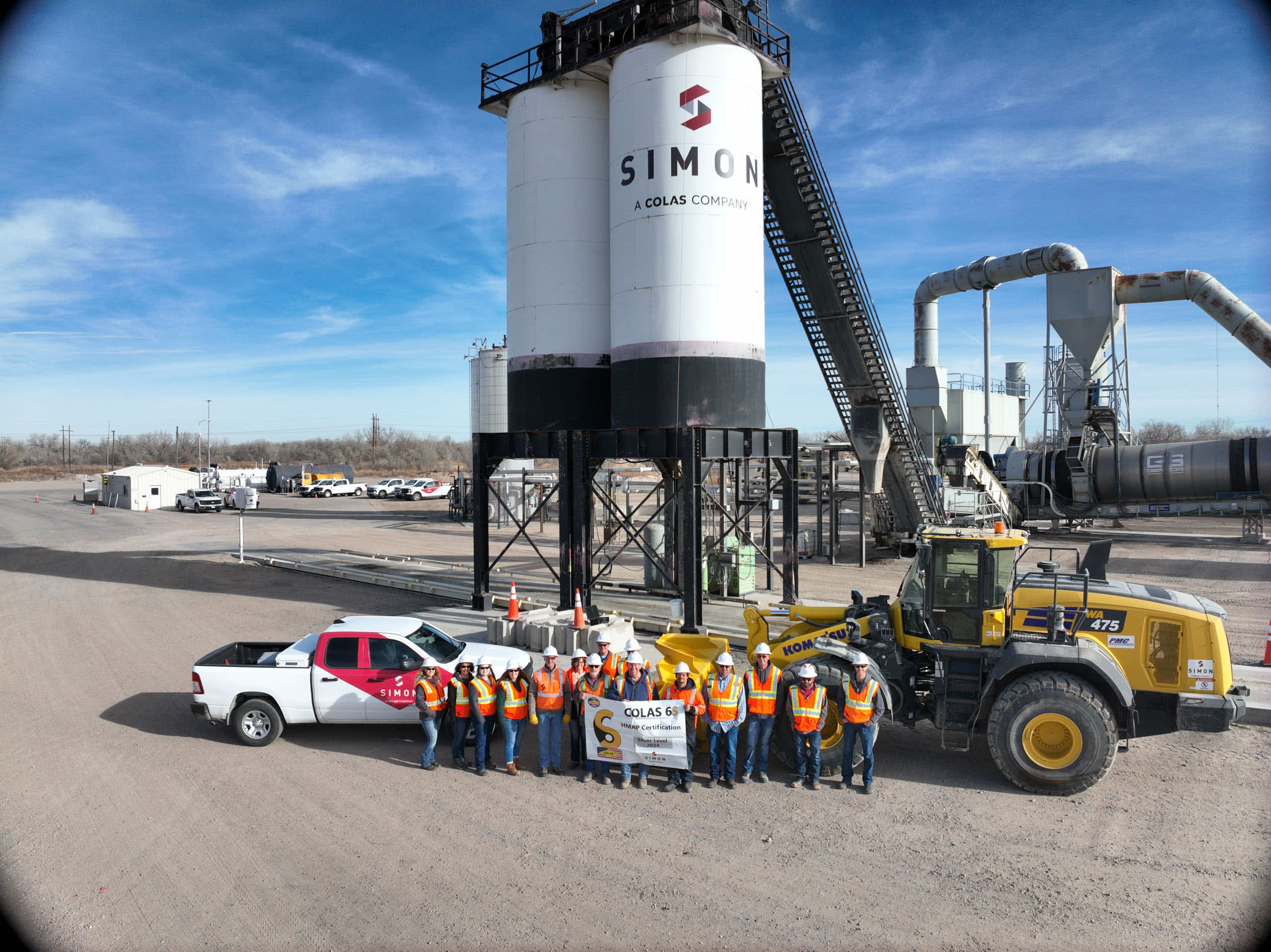 Group photo of construction workers in safety gear standing in front of Simon Company machinery and tanks at a job site.