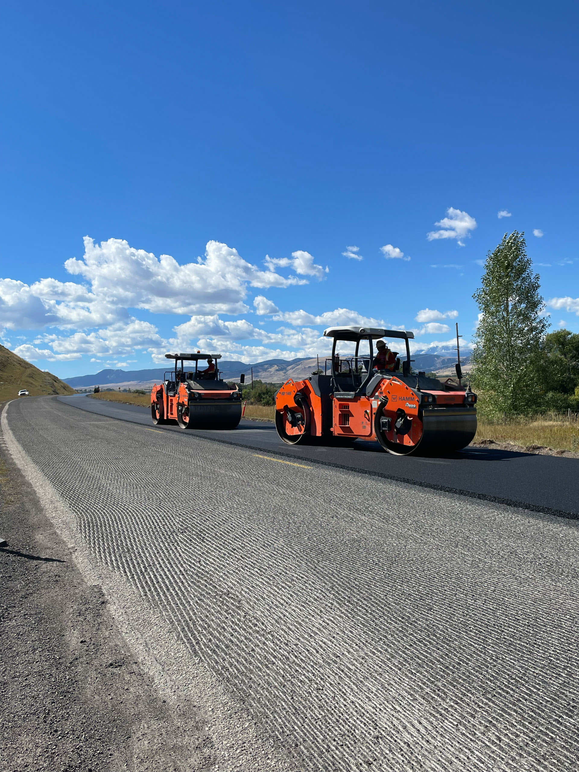Two road rollers working on a freshly paved road under a blue sky with clouds.