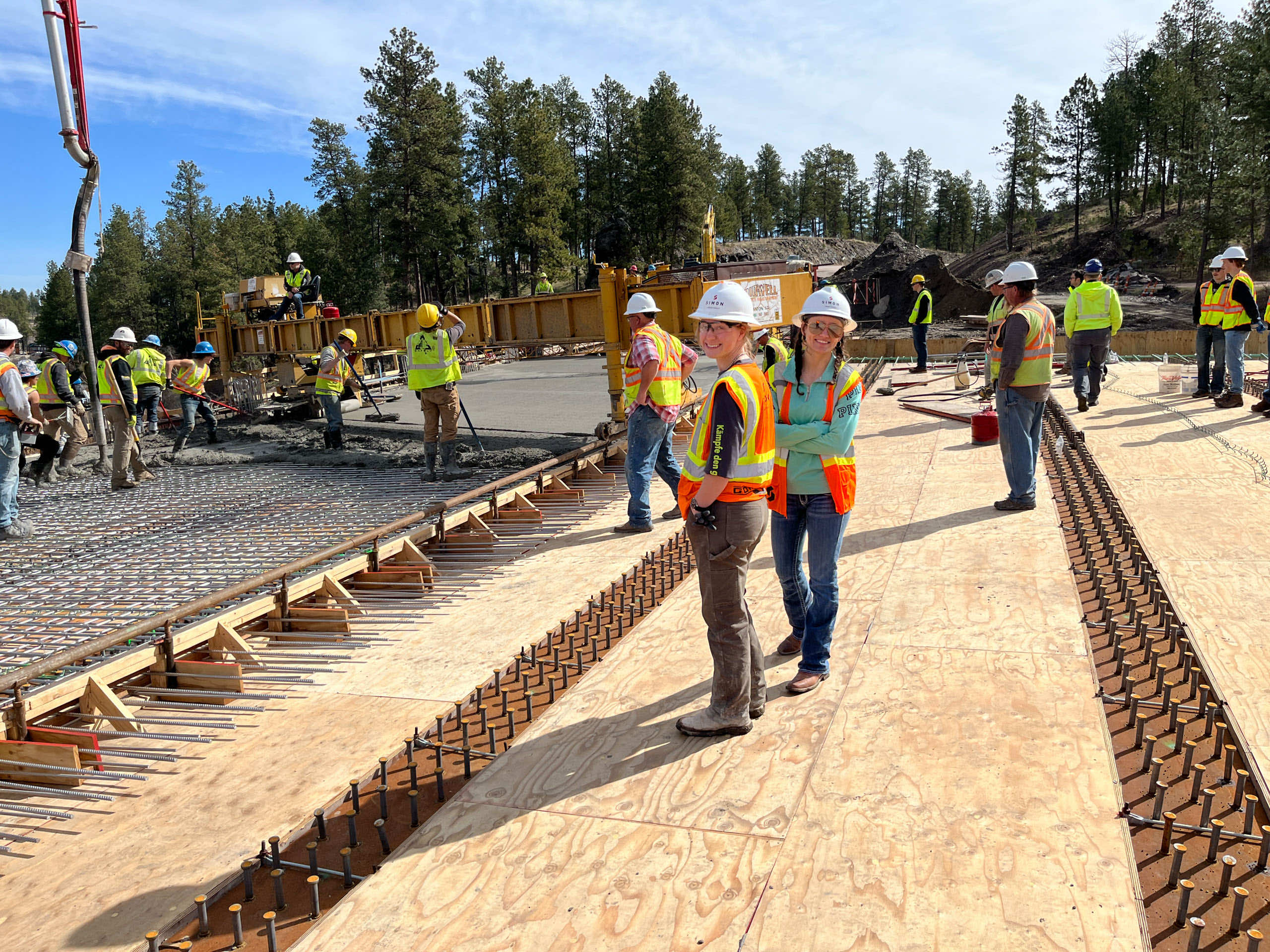 Construction workers on a site pouring concrete with equipment and trees in the background.