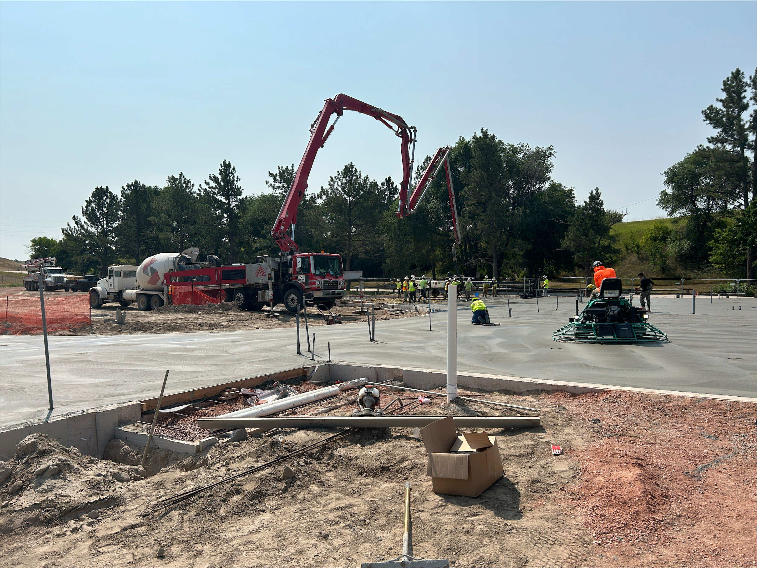 Construction site with workers pouring concrete for a foundation using a concrete pump and various machinery.